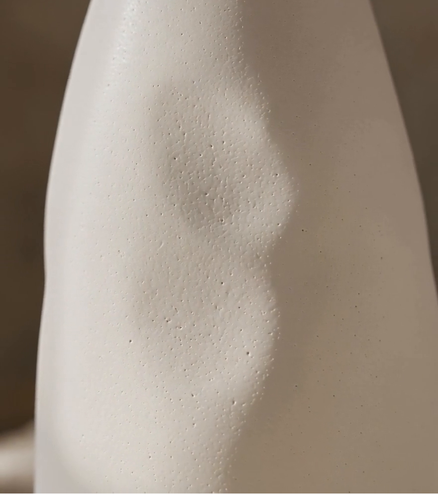 A close-up of a matte off-white ceramic vase with a subtle textured surface and soft shadow details, set against a warm, neutral background.