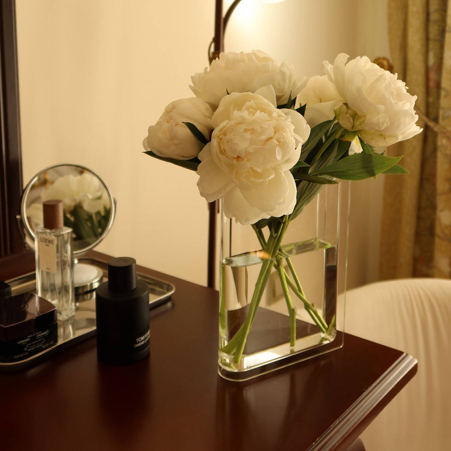 Le Vase Livre with white peonies on a dark wood vanity beside perfume bottles and a round mirror